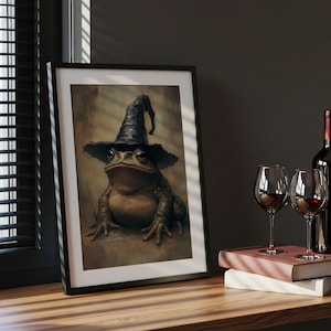 May include: Framed artwork depicting a frog wearing a black witch's hat. The frog is rendered in detail with brown and green skin. The artwork is displayed on a wooden surface, with wine glasses and a bottle of wine in the background.