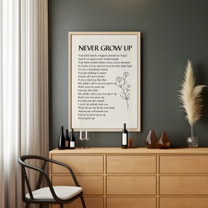 May include: A framed print with a white background and black text. The text reads "NEVER GROW UP" and includes a poem about staying young at heart. The print is hanging on a wall above a wooden cabinet with wine bottles and glasses.