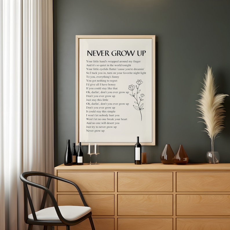 May include: A framed print with a white background and black text. The text reads "NEVER GROW UP" and includes a poem about staying young at heart. The print is hanging on a wall above a wooden cabinet with wine bottles and glasses.