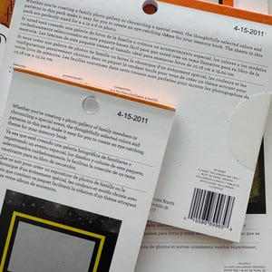 K&ouml;nnte beinhalten: Zwei gestapelte Packungen mit dekorativen Papierb&ouml;gen f&uuml;r Scrapbooking, mit orangefarbenen und wei&szlig;en Kopfzeilen und dem Datum 4-15-2011. Die Verpackung enth&auml;lt mehrsprachige Beschreibungen, ein kreisf&ouml;rmiges Aufh&auml;ngeloch und einen Strichcode.