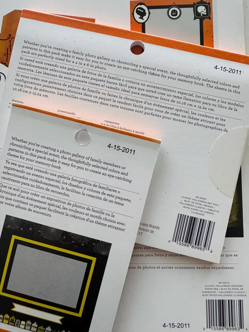 K&ouml;nnte beinhalten: Zwei gestapelte Packungen mit dekorativen Papierb&ouml;gen f&uuml;r Scrapbooking, mit orangefarbenen und wei&szlig;en Kopfzeilen und dem Datum 4-15-2011. Die Verpackung enth&auml;lt mehrsprachige Beschreibungen, ein kreisf&ouml;rmiges Aufh&auml;ngeloch und einen Strichcode.