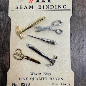 May include: A vintage card featuring miniature sewing tools: two hammers, two scissors, and a needle tool. The card reads "Warren's SEAM BINDING" and "FINE QUALITY RAYON". The tools are gold and silver-toned.