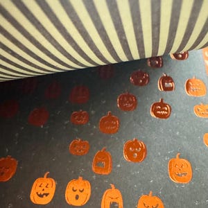 K&ouml;nnte beinhalten: Ein Blatt dunkelgraues Papier mit einem sich wiederholenden Muster aus kleinen, metallisch orangefarbenen K&uuml;rbissen mit verschiedenen Gesichtsausdr&uuml;cken. Der obere Teil des Bildes zeigt eine gefaltete Kante mit hellgelb-dunkelgrauem Streifenmuster.