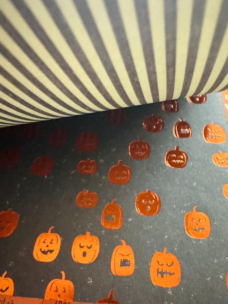K&ouml;nnte beinhalten: Ein Blatt dunkelgraues Papier mit einem sich wiederholenden Muster aus kleinen, metallisch orangefarbenen K&uuml;rbissen mit verschiedenen Gesichtsausdr&uuml;cken. Der obere Teil des Bildes zeigt eine gefaltete Kante mit hellgelb-dunkelgrauem Streifenmuster.