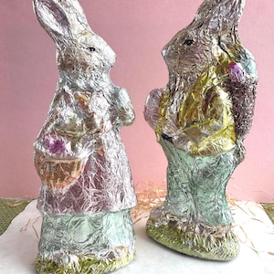May include: Two foil-wrapped Easter bunny figures. One bunny is wearing a pink dress and holding a basket with a purple egg. The other bunny is wearing a yellow shirt and green pants and holding a basket with a pink egg.