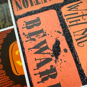 K&ouml;nnte beinhalten: Eine dekorative Collage mit rechteckigen orangefarbenen Feldern, schwarzem Text mit der Aufschrift CAUTION und BEWARE sowie einem schwarzen Glitzerrand. Das Design umfasst eine schwarze Glitzer-Kr&auml;hensilhouette und ein K&uuml;rbis-Motiv.