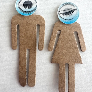 May include: Two brown cardboard figures, one male and one female, each with a circular blue and white eye design for a head. The male figure is a simple cutout, while the female figure has a skirt. The background is white.
