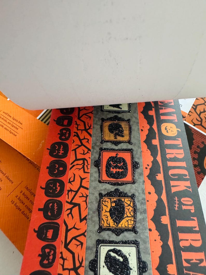 K&ouml;nnte beinhalten: Ein dekorativer Papierblock mit Halloween-Motiven in Orange und Schwarz. Die Seiten zeigen Muster von K&uuml;rbissen, kahlen &Auml;sten und gerahmten Symbolen von Totenk&ouml;pfen und Raben. Der Text EAT TRICK OR TREAT ist vertikal aufgedruckt.