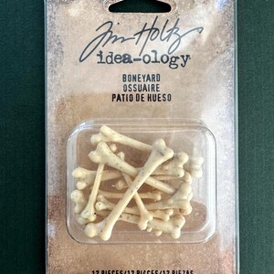 May include: A package of 12 small, off-white bone-shaped pieces with dark speckles, labeled Tim Holtz idea-ology Boneyard. The items are enclosed in a clear plastic blister pack attached to a brown, weathered-looking cardboard backing card.