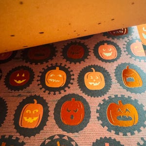 K&ouml;nnte beinhalten: Ein gemustertes Papierblatt mit einem sich wiederholenden Design aus metallisch orangefarbenen K&uuml;rbissen, die jeweils von einem dunklen, gewellten kreisf&ouml;rmigen Rand umschlossen sind. Der Hintergrund besteht aus einer texturierten, violetten Fl&auml;che.