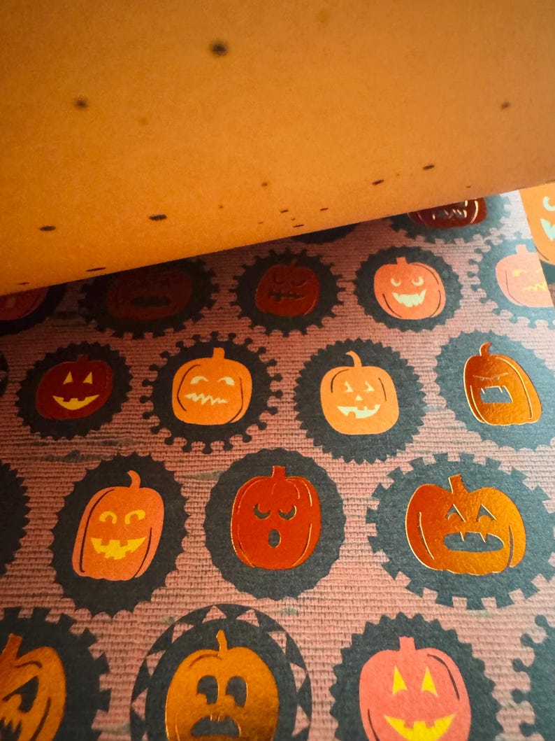 K&ouml;nnte beinhalten: Ein gemustertes Papierblatt mit einem sich wiederholenden Design aus metallisch orangefarbenen K&uuml;rbissen, die jeweils von einem dunklen, gewellten kreisf&ouml;rmigen Rand umschlossen sind. Der Hintergrund besteht aus einer texturierten, violetten Fl&auml;che.