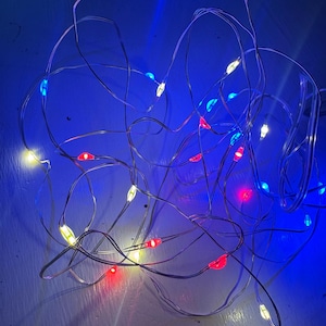 May include: A tangled string of fairy lights with small, colorful LED bulbs. The lights are a mix of red, blue, yellow, and white, creating a festive and vibrant display. The thin, silver wire is visible, and the lights are scattered against a blue background.