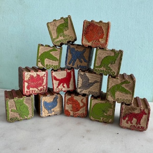 May include: A collection of vintage wooden blocks arranged in a pyramid. Each block features a raised, colored relief illustration of an animal or object, including a kangaroo, Indian, goose, lamb, fox, duck, nest, owl, hen, mouse, cat, and jackal.