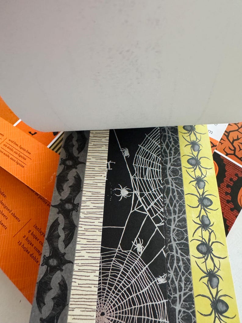 K&ouml;nnte beinhalten: Ein Papierblock mit verschiedenen Halloween-Mustern, darunter schwarze Flederm&auml;use auf grauem Grund, eine wei&szlig;e Birkenrindenstruktur, wei&szlig;e Spinnennetze auf schwarzem Grund, graue Baumzweige und schwarze Spinnen auf gelbem Grund.