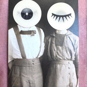 May include: A black and white collage print featuring two figures with circular paper eyes replacing their heads. The left figure wears a collared shirt with a bow tie and suspenders, while the right figure wears a buttoned shirt. The eyes are white with black pupils and long lashes.