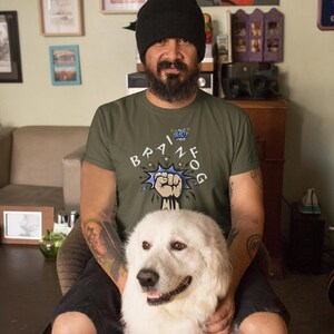 May include: An olive green t-shirt with the text "BRAIN FOG" in a comic book style. A white Great Pyrenees dog is in the foreground. The person in the image is wearing a black beanie.