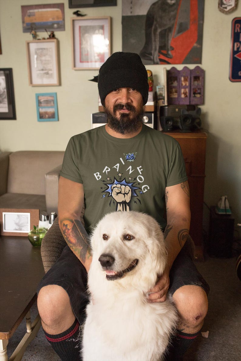 May include: An olive green t-shirt with the text "BRAIN FOG" in a comic book style. A white Great Pyrenees dog is in the foreground. The person in the image is wearing a black beanie.