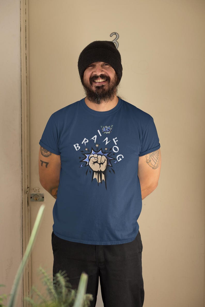 May include: Navy blue t-shirt with the text "BRAIN FOG" in a comic book style. The design includes a clenched fist and stars. The person in the image is wearing a black beanie and black pants.