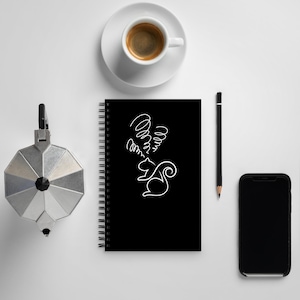 May include: A black spiral-bound notebook with a white line drawing of a squirrel and swirling lines. A cup of coffee, a pencil, a phone, and a silver coffee maker are also on the white surface.