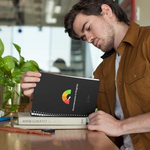 May include: A black spiral-bound notebook with the text "Decompression in Progress" and a colorful graphic. The notebook is resting on a stack of books, with a green pen and pencils nearby. A potted plant is in the background.
