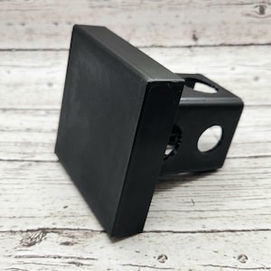 以下が含まれることがあります： 黒い金属製のヒッチレシーバーに、正方形の黒いプラスチック製のカバーが付いています。レシーバーには、側面に2つの穴と、上部に1つの丸い穴があります。