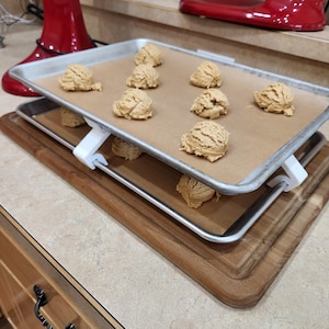 May include: Two metal baking sheets with parchment paper, stacked with white supports, holding scoops of cookie dough. The sheets rest on a wooden cutting board. A red stand mixer is visible in the background.
