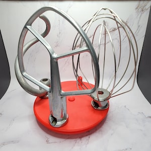 May include: A set of stainless steel mixer attachments, including a dough hook and a wire whisk, resting on a red base. The attachments are designed for a stand mixer, ideal for baking and cooking.