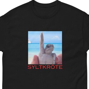Op de afbeelding: Een zwart t-shirt met korte mouwen en een vierkante grafische print op de borst. De print toont een kleine zeeschildpad op een strand met de oceaan op de achtergrond, en de rode tekst SYLTKRÖTE direct onder de afbeelding gedrukt.