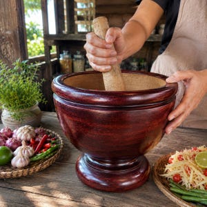 May include: A large, dark reddish-brown wooden mortar and pestle set. The mortar features a smooth, rounded bowl shape with a pedestal base, while the pestle is a cylindrical wooden tool used for grinding ingredients within the bowl.