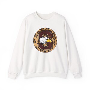 May include: White sweatshirt featuring a circular design with a leopard print background and the letter "S" in a maroon color. An eagle head with white feathers and a yellow beak is inside the letter.