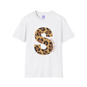 May include: A white short-sleeved t-shirt featuring a large letter S with a brown and black leopard print pattern. The text SAYRE EAGLES is printed in a vertical brown font along the right side of the letter S. A small blue logo is on the inner neck.