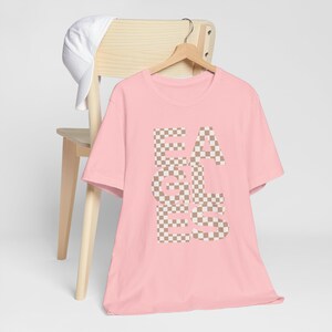 Könnte beinhalten: Hellrosa T-Shirt mit dem Wort "EAGLES" in einem karierten Muster aus Beige und Off-White. Eine weiße Baseballkappe liegt auf einem Holzstuhl hinter dem Shirt.