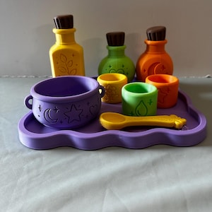 May include: A set of colorful toy potion bottles, small cups, a purple cauldron, and a yellow spoon arranged on a wavy purple tray. The items feature embossed designs including a moon, stars, hearts, leaves, flowers, and a water droplet.