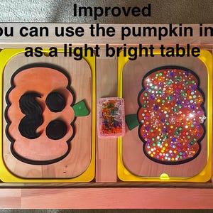 May include: A wooden table with two pumpkin-shaped inserts. The left insert has black facial features, while the right insert is illuminated with colorful lights. A small container of translucent pegs sits between them. Text reads: Improved you can use the pumpkin insert as a light bright table.