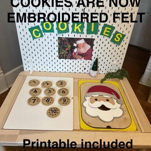 May include: A wooden table displays a white pegboard with the text "IMPROVED COOKIES ARE NOW EMBROIDERED FELT COOKIES." A Santa Claus image is attached. The table also holds numbered cookie-shaped felt pieces and a Santa Claus puzzle.
