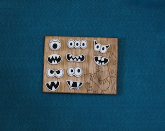 Puede incluir: Una tabla de madera con varios recortes de caras de monstruos. Los recortes incluyen ojos, dientes y cuernos. La tabla es de color marrón claro y tiene una veta de madera natural.