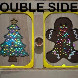 May include: Double-sided wooden light-up board with a Christmas tree and a gingerbread man. The board has a yellow frame and is lit up with colorful lights.