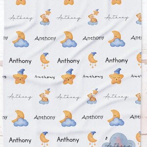 Peut inclure: Une couverture de bébé blanche avec un motif répété d'étoiles, de lunes et de nuages. Le nom "Anthony" est répété tout au long du motif.