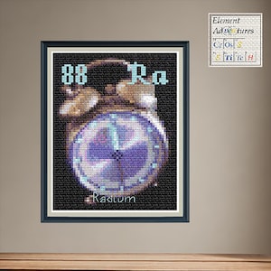 May include: Framed cross-stitch art featuring a vintage-style clock design. The clock face is a gradient of purple and blue, with the word "Radium" below. The top of the frame has the numbers "88" and the letters "Ra". The artwork is displayed on a neutral wall.