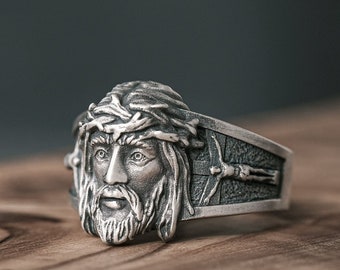 May include: A silver ring with a detailed depiction of Jesus Christ's face, wearing a crown of thorns. The ring band features a cross design.