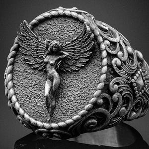 Peut inclure: Bague argentée détaillée représentant une figure ailée sculptée dans un cadre ovale texturé. La bande de la bague est ornée de volutes complexes et d'une bordure tressée, mettant en valeur un design classique.