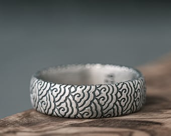 May include: A silver ring with a detailed, intricate pattern of swirling lines. The ring is on a wooden surface.