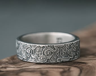 May include: A silver ring with a textured design of repeating floral patterns. The ring is sitting on a piece of wood.