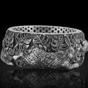 May include: A silver-colored ring with a detailed design of a rabbit resting among leaves. The ring has a wide band with a textured surface and a series of small holes around the top edge. The rabbit and leaf design is intricately carved.
