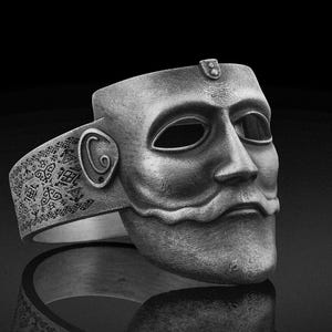 Puede incluir: Un anillo de tono plateado que presenta un diseño de máscara de rostro humano esculpido. La banda está decorada con intrincados patrones geométricos pequeños y símbolos. La máscara tiene ojos, nariz y bigote definidos, con un detalle circular.