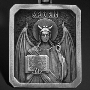 May include: A silver pendant necklace with a square-shaped charm featuring a detailed depiction of Satan with wings and a book. The charm has the word "Satan" inscribed at the top.