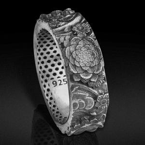 May include: A silver ring with intricate floral carvings. The ring features a large, detailed flower design on the outer band, with smaller floral accents and a textured interior. The ring is marked with "925".