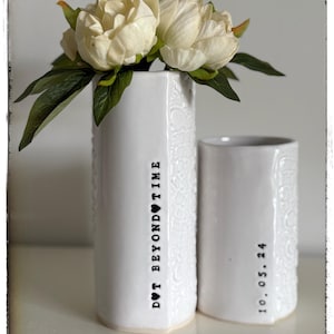 May include: Two white ceramic vases with embossed designs. The vase on the left has the text "Don't Beyond Time" with a heart symbol. The vase on the right has the date "10.05.24" inscribed on it. A bouquet of white peonies is in the vase on the left.