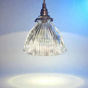 Op de afbeelding: Een heldere glazen hanglamp met een gegolfde, klokvormige kap. De lamp heeft een bronzen metalen basis en een enkele lamp. De kap heeft verticale ribbels, waardoor een textuureffect ontstaat. De lamp hangt aan een draad.