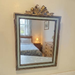 Op de afbeelding: Rechthoekige spiegel met een goudkleurige strik aan de bovenkant. De lijst is goudkleurig en lichtblauw-groen. De spiegel reflecteert een slaapkamer met een bed, nachtkastje en dressoir.